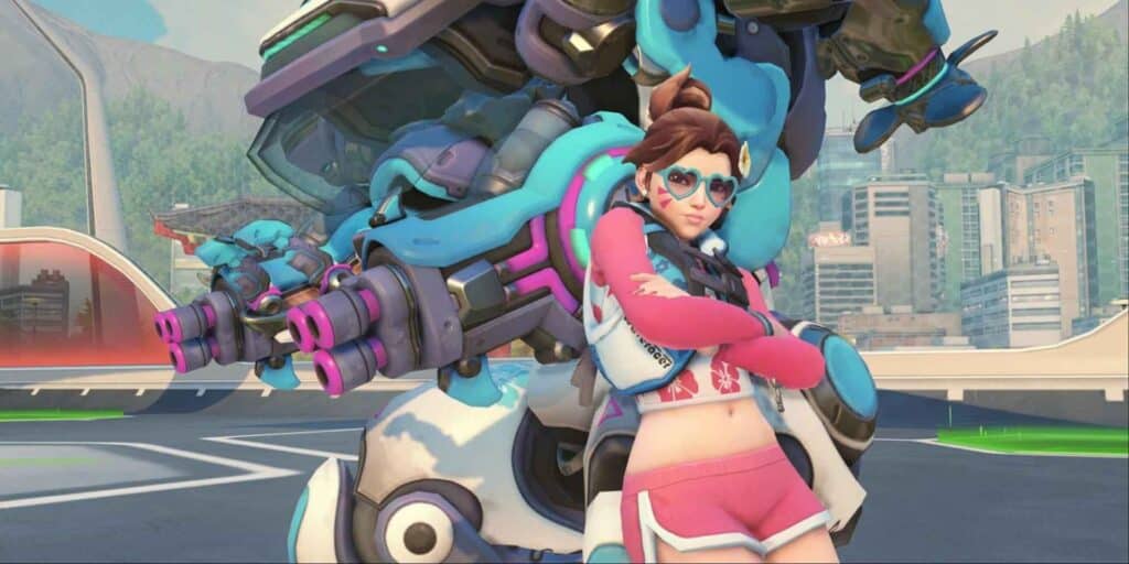 Overwatch 2 character with heart-shaped glasses and pink attire stands confidently with arms crossed in front of a large blue and pink mech, cityscape in background.