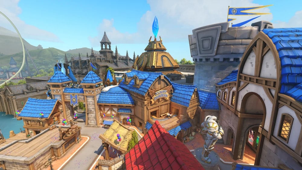 A vibrant, fantasy-themed town with blue-roofed buildings, a castle tower, balloons, a statue, and banners. The scene is lively and whimsical
